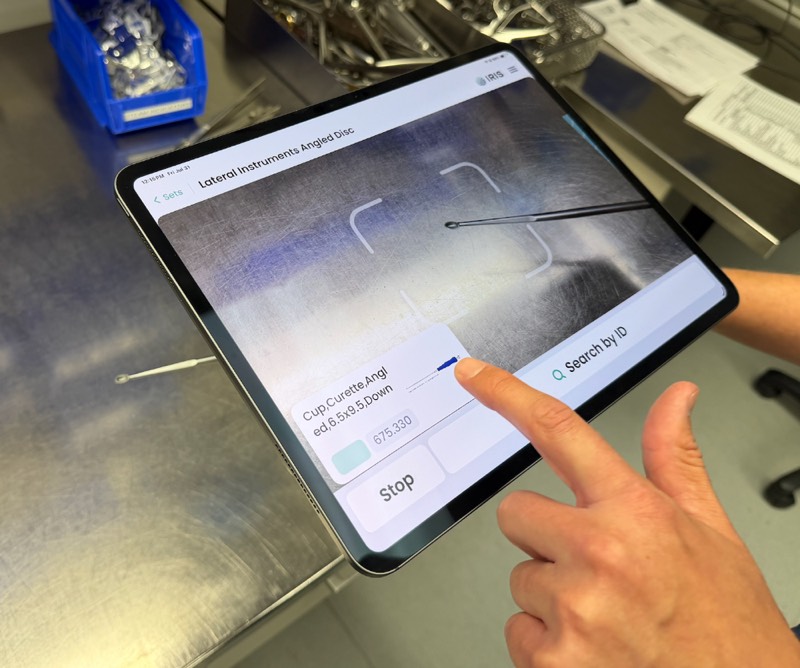 A hand using the IRIS app on a tablet over a tray of instruments